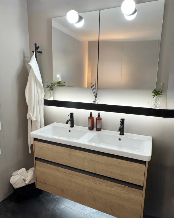 Double white sinks on a wooden floating vanity with black taps, a two-panel mirror, globe lights, a robe on a hook, towels on the floor, and minimal plant decor.