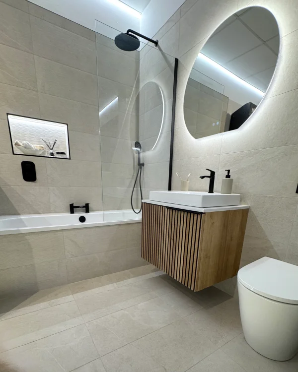 Modern bathroom with a white bathtub, glass shower panel, black fixtures, wall-mounted wooden slatted vanity with a square sink, and a large round backlit mirror.