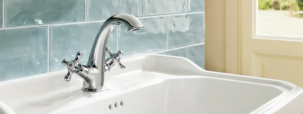 Chrome bathroom faucet with cross handles above a white sink, with subway tile backsplash and window in background.