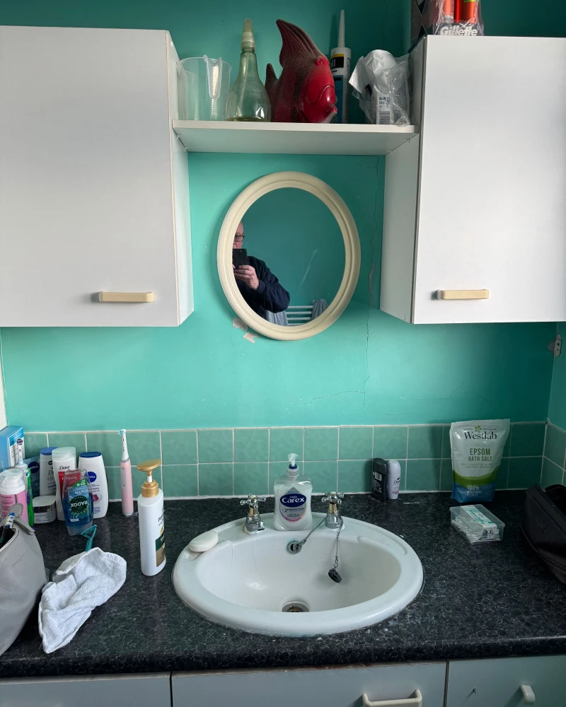 Teal bathroom with white cabinets and a round mirror; a person takes a photo in the mirror. A red fish ornament and bottles sit on the shelf above the sink.