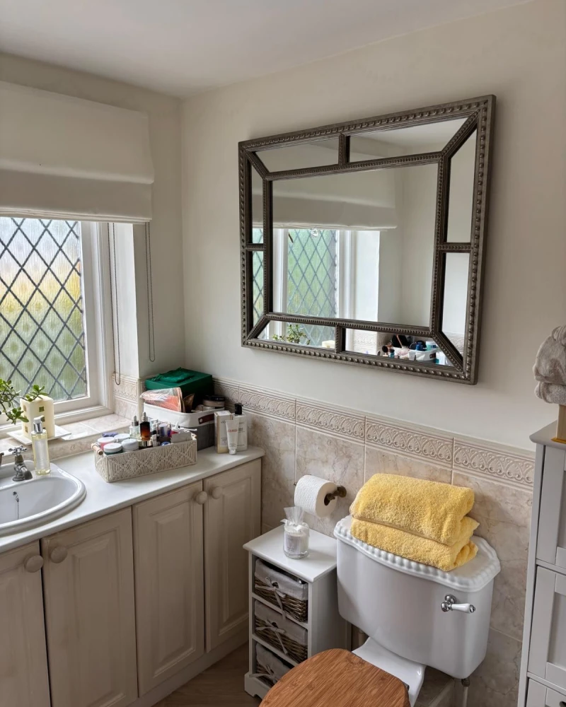 Bright bathroom with cream cabinetry, beige tiled walls, a large ornate rectangular mirror, a lattice-pane window, and a toilet topped with yellow towels.