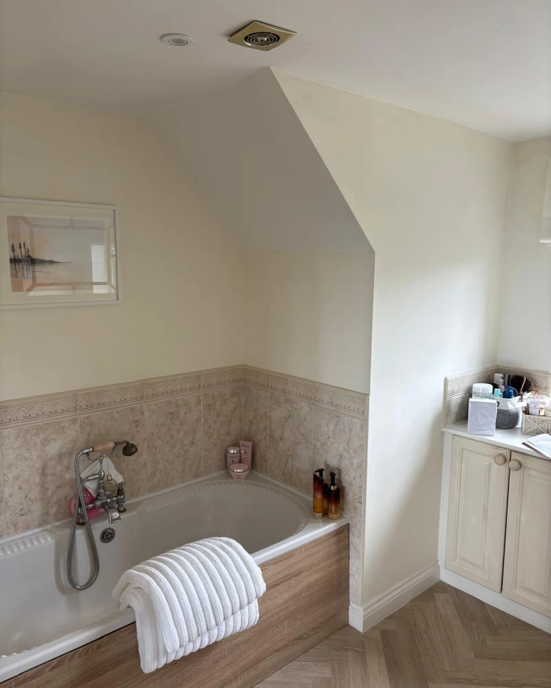 Bright bathroom with a built-in tub in wood paneling, white towel draped over edge, beige tiled surround, framed seascape and a cabinet with toiletries.