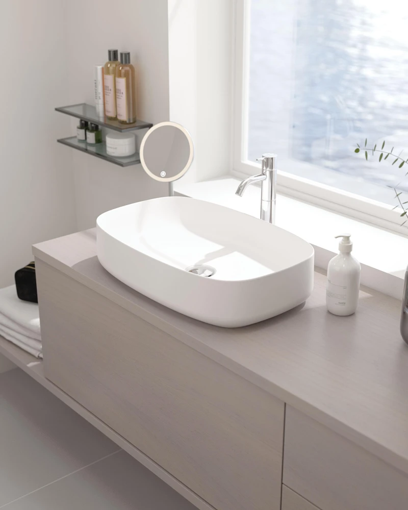 Modern bathroom with a white oval vessel sink on a light wood vanity, chrome faucet, round mirror, glass shelves with bottles, and a window by the water.