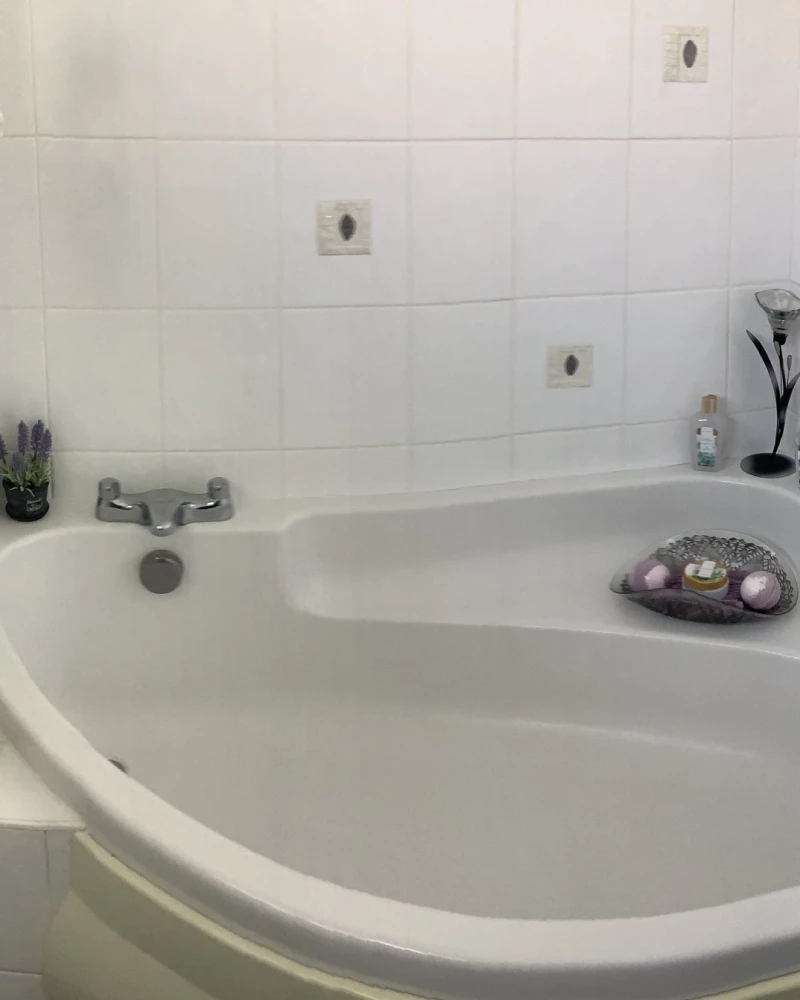 White tiled bathroom with a curved corner tub; left features a faux flowering tree and lavender, with bath products on ledge and a tray of bath bombs.
