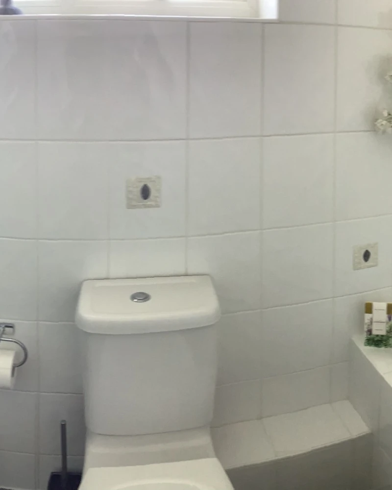 White-tiled bathroom with sink left, purple towel on a rail, toilet centre, tub right, backpack on the floor, toiletries on the tub ledge.