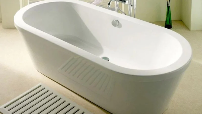 Modern white freestanding bathtub in a bright bathroom, chrome tap, a slatted floor vent in the foreground, and a green plant in a vase nearby.