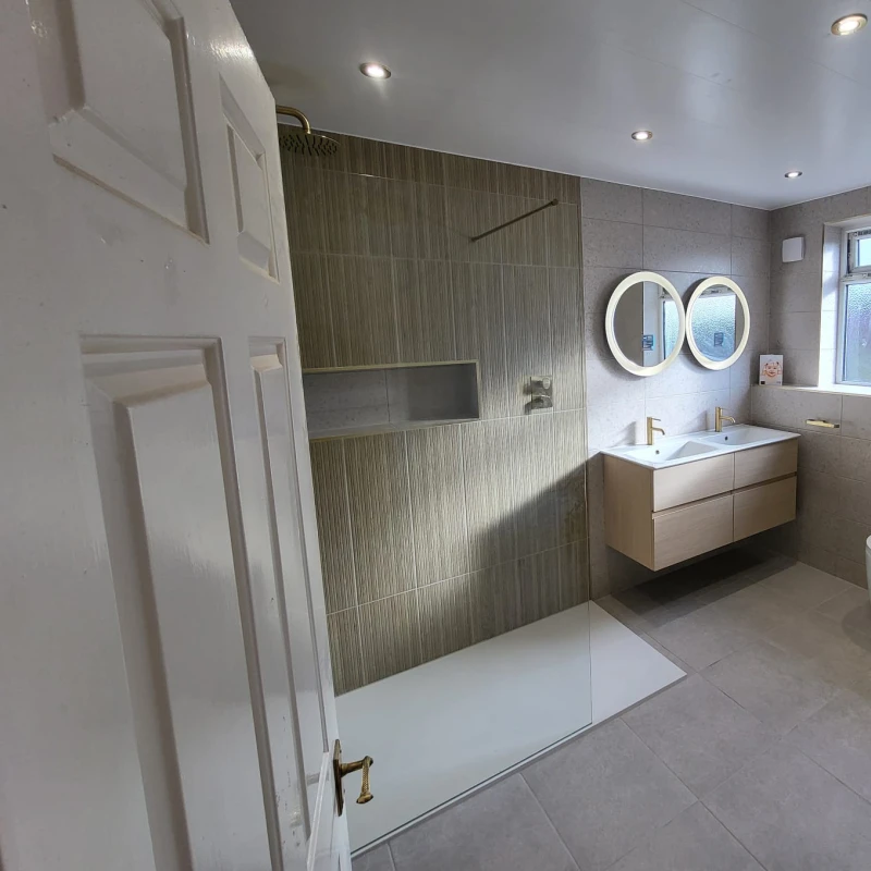 Contemporary bathroom with a glass shower enclosure, grey tiled walls, a wood-tone double vanity with brass taps, and two round backlit mirrors.
