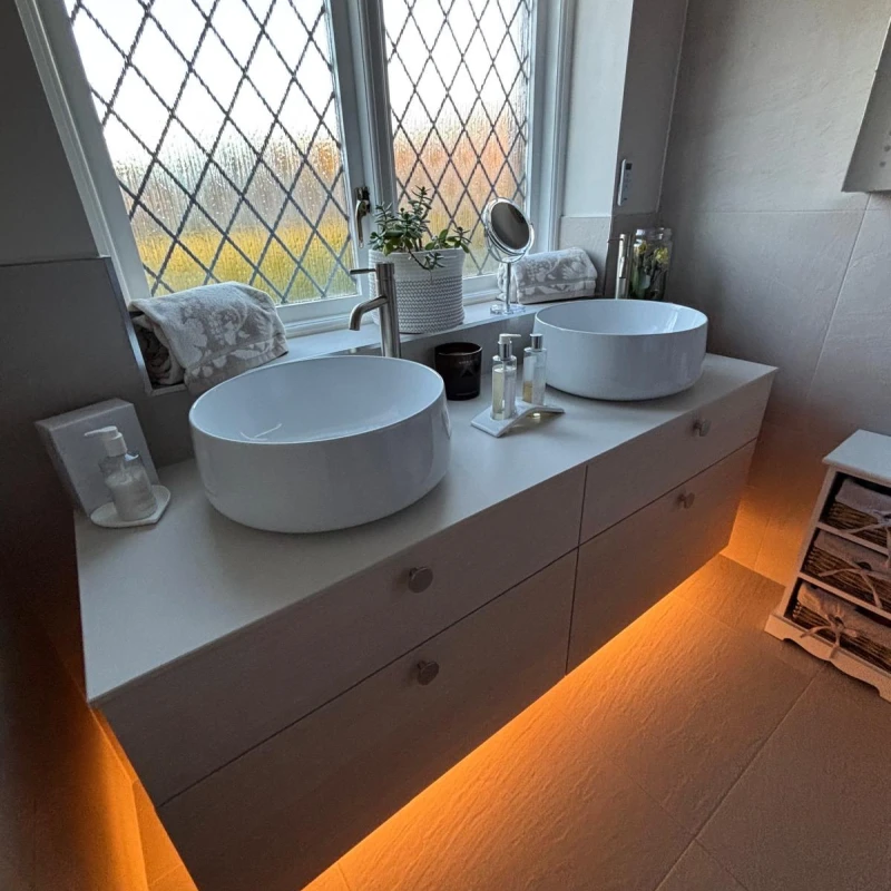 Modern bathroom with two white vessel sinks on a white vanity, diamond-pattern frosted window, warm LED under-cabinet lighting, towels and a plant.