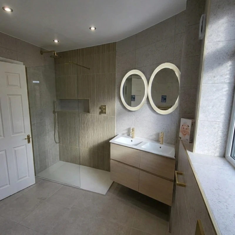 Modern bathroom with a glass-walled shower, double sink vanity, two round mirrors, grey tiled walls and floor, and a frosted window.