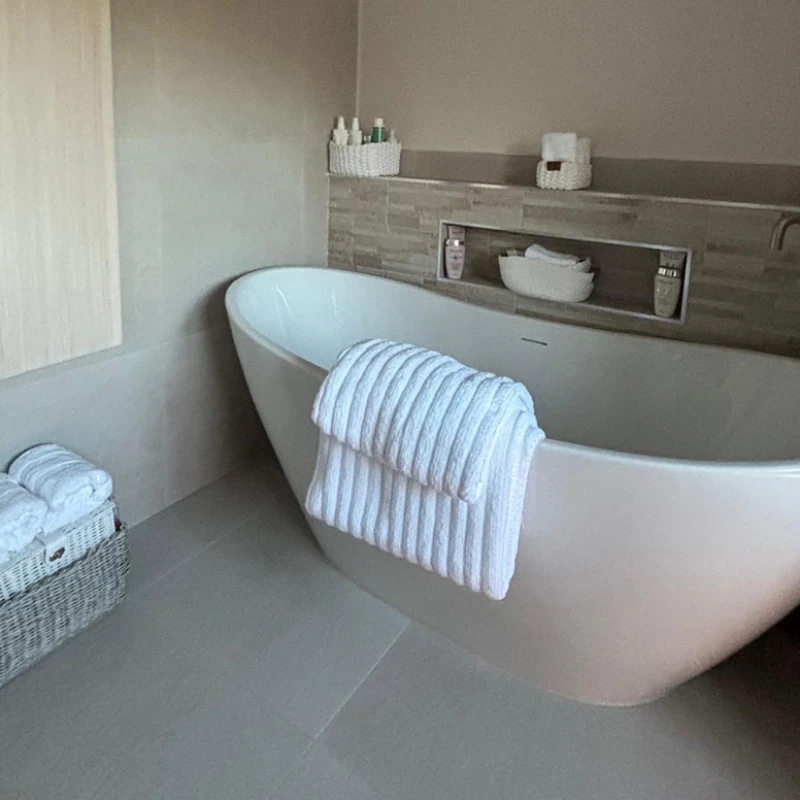 Modern bathroom with a white freestanding tub, folded towel draped over the rim, tiled wall with a built-in shelf, and baskets of towels and toiletries.