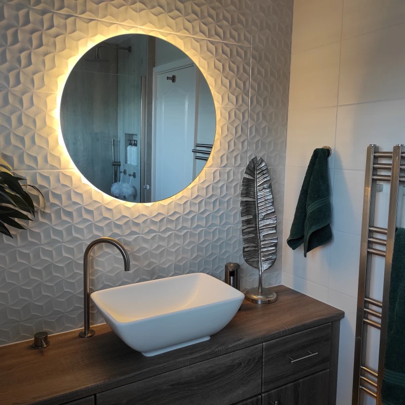 Round backlit mirror above white vessel sink on wooden vanity, textured wall tiles, plant, metal leaf sculpture, and green towels on a chrome warmer.