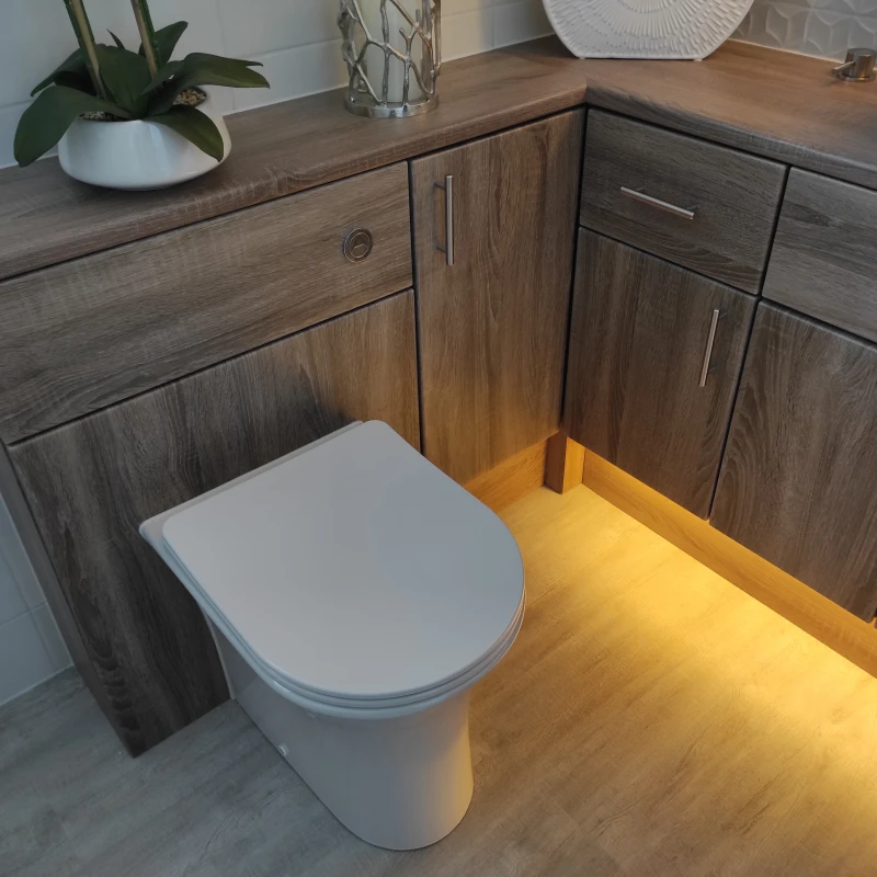 Corner bathroom with dark woodgrain cabinetry around a white toilet; warm under-cabinet lighting, a plant on counter and a white sink at right.