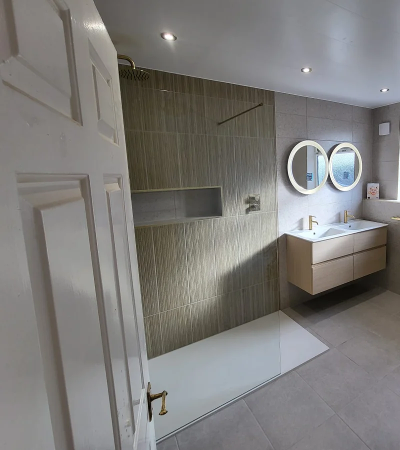 Contemporary bathroom with a glass shower enclosure, grey tiled walls, a wood-tone double vanity with brass taps, and two round backlit mirrors.