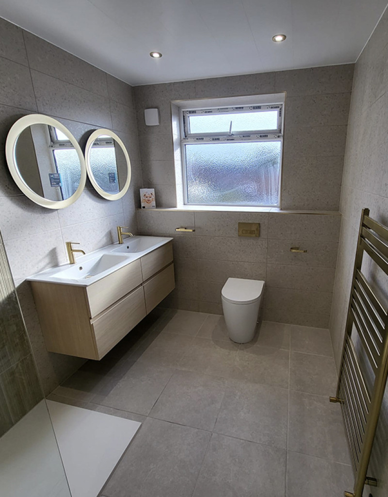 Modern bathroom with a double-sink vanity, round backlit mirrors, frosted window, glass shower on the left, gold fixtures, a freestanding toilet and brass towel rail.