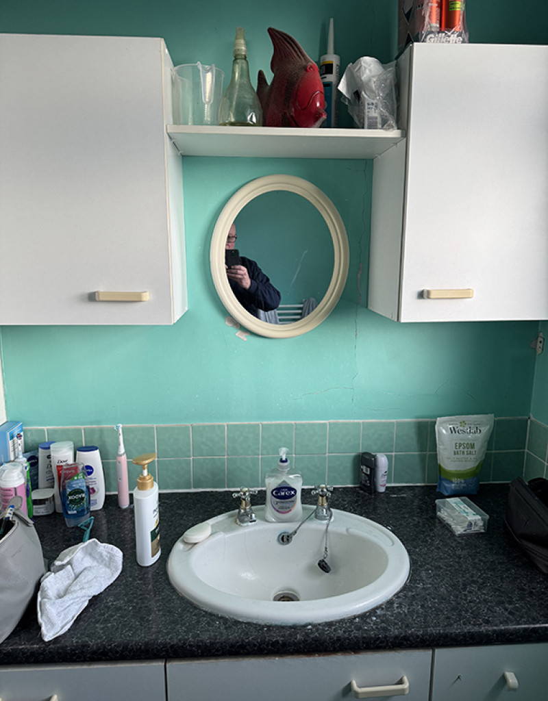 Bathroom vanity with white cabinets, round mirror and teal wall. Black granite countertop with sink, soap, and toiletries; person reflected taking the photo.