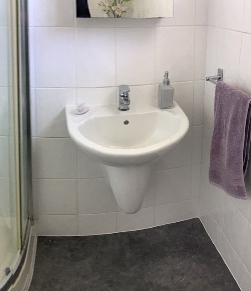 Small white wall-mounted pedestal sink in a tiled bathroom, chrome tap, soap dispenser, purple towel on a rail, and curved glass shower to the left.
