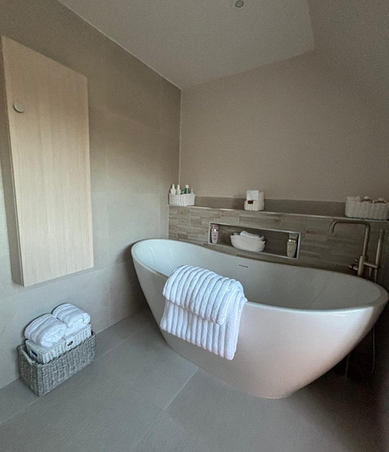 A modern bathroom with a white freestanding tub, a folded towel over the edge, grey walls, and a wicker basket of towels beside the tub.