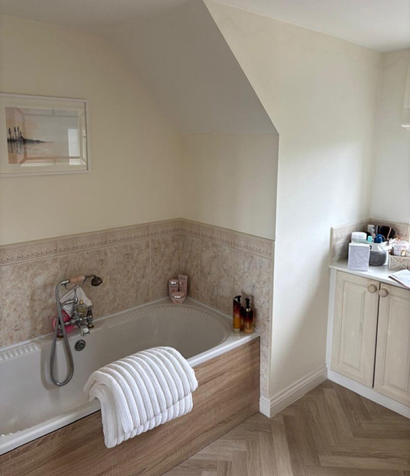 Bright bathroom with a built‑in bathtub along a tiled wall, a white towel draped over the edge, handheld shower, and cream cabinets with toiletries on the right.